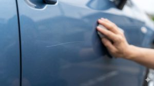 査定前に車のバンパーの擦り傷を確認し、修理するか悩んでいる日本人男性。背景にはガレージとシルバーの乗用車。